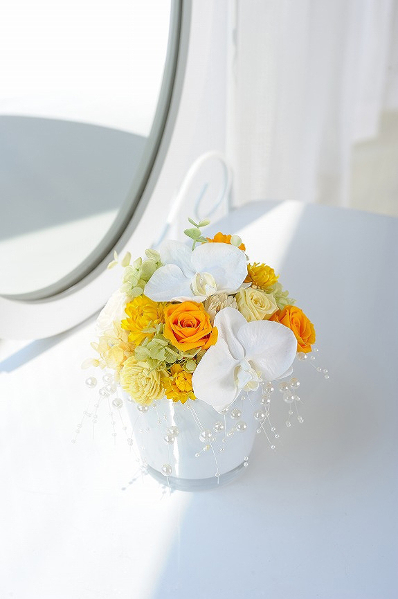 <p>薄いグレーのガラス鉢に仕立てられた暖色系プリザに白の胡蝶蘭。</p>