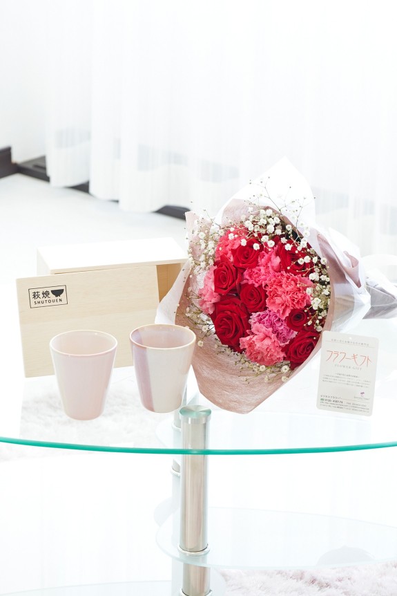 母の日の定番カーネーション＆皇室献上、農林水産大臣賞の受賞実績のあるバラ農園のバラをミックスさせた花束・ブーケに「いつもありがとう」の3D立体ピックを挿したお花と、すっきりとしてスマートなデザインで老若男女を問わない萩焼のペアカップをセットにしたフラワーギフト用の商品です。