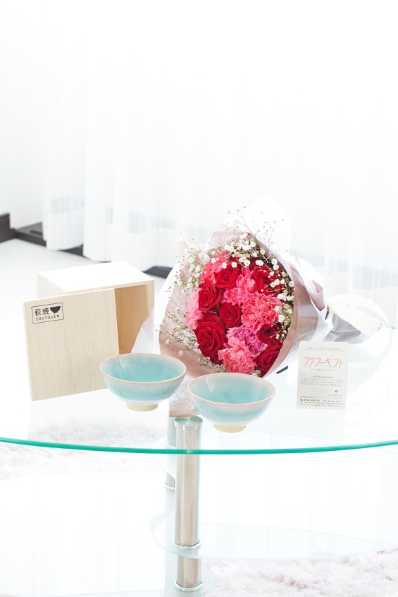 母の日の定番カーネーション＆皇室献上、農林水産大臣賞の受賞実績のあるバラ農園のバラをミックスさせた花束・ブーケに「いつもありがとう」の3D立体ピックを挿したお花と、すっきりとしてスマートなデザインで老若男女を問わない萩焼のペアお茶碗をセットにしたフラワーギフト用の商品です。